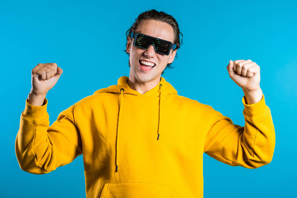 Hipster man in glowing glasses moving to the music rhythm on blue studio background. Dancing positive comic guy - Foto, Imagem