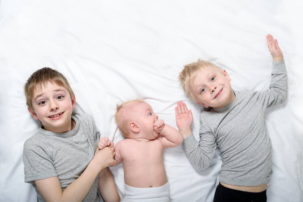 Zwei ältere Brüder liegen mit dem jüngsten Baby in einem weißen Bett. glückliche Kindheit, große Familie - Foto, Bild