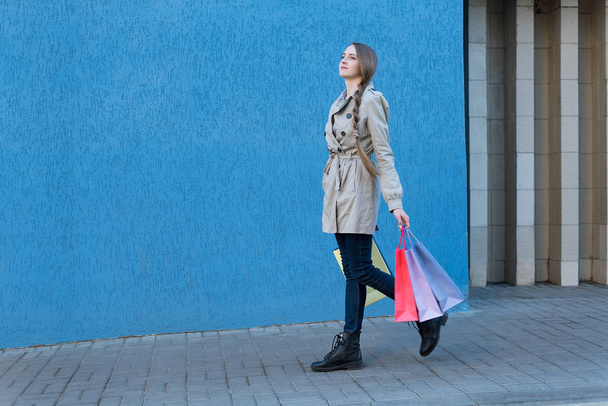 Renkli çantaları olan güzel genç bir kadın sokakta yürüyor. Arkaplanda mavi duvar - Fotoğraf, Görsel