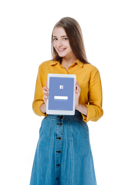 KYIV, UKRAINE - AUGUST 12, 2019: smiling girl in denim skirt holding digital tablet with Facebook app isolated on white - 写真・画像
