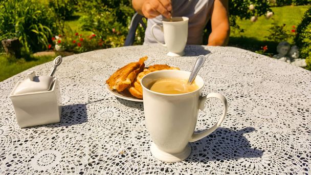 Terveellinen aamiainen vihreässä puutarhassa kesäaikaan. Tarjoillaan kahden kupillisen kahvia maidon ja kuumien voileipien kanssa. Mies istuu vastapäätä, aamiainen kahdelle
. - Valokuva, kuva