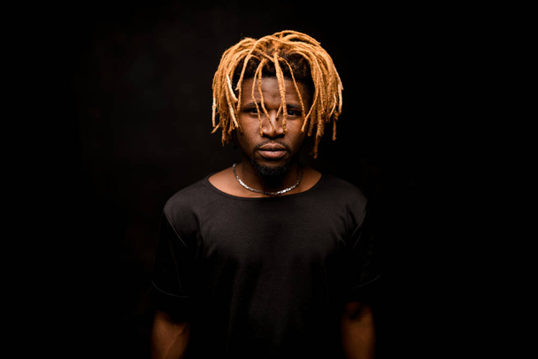 Black guy with blonde dreadlocks posing in the dark outfit looking at camera - Photo, Image
