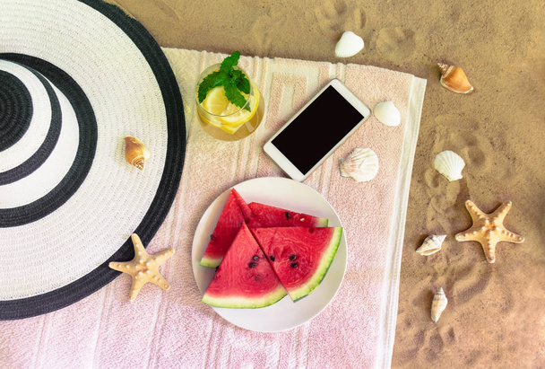 Gran sombrero de playa, smartphone, vaso de limonada y plato de sandía sobre toalla en playa de arena
.  - Foto, Imagen