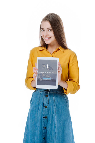 KYIV, UKRAINE - AUGUST 12, 2019: smiling girl in denim skirt holding digital tablet with tumblr app isolated on white - 写真・画像