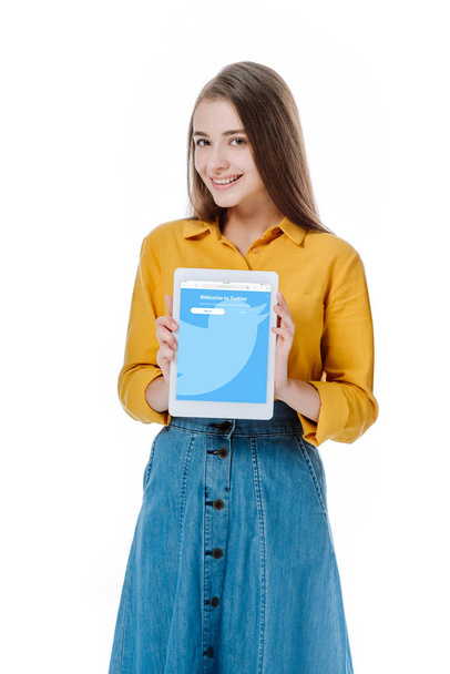 KYIV, UKRAINE - AUGUST 12, 2019: smiling girl in denim skirt holding digital tablet with twitter app isolated on white - 写真・画像