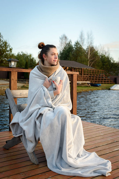 Egy fiatal lány egy meleg kockás ruhában fekszik a tónál ősszel. A természet élvezetének, kikapcsolódásnak, utazásnak a koncepciója - Fotó, kép