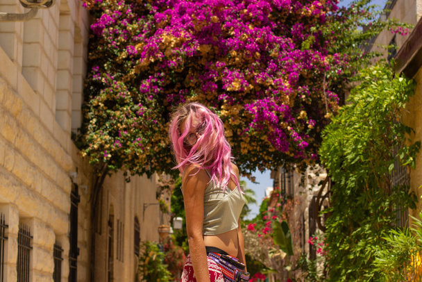 jong meisje portret met gezicht achter roze haar in het centrum van de compositie op de oude stad straat met roos en groene plant en bloemen achtergrond in zonnige kleurrijke weer tijd  - Foto, afbeelding