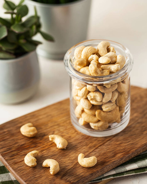 A lot of cashew nuts in a glass container on a wooden Board on a light background. In the background, green plants in pots. - Photo, Image