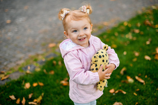 Улыбающаяся маленькая девочка, стоящая в осеннем парке и обнимающая жёлтую чёрную точеную игрушку
 - Фото, изображение