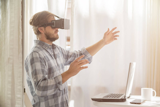 Homem barbudo jovem vestindo óculos de realidade virtual no interior moderno. Bonito homem caucasiano usando fone de ouvido VR, jogando videogames
. - Foto, Imagem