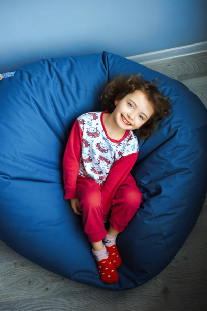 Niño de 6 años de edad de ojos azules niña en pijama rosa y calcetines rojos yacen en otomana azul y mira hacia arriba con sonrisa. día de sueño. Felicidad, ocio y estilo de vida en la infancia. Luz natural del día
 - Foto, imagen