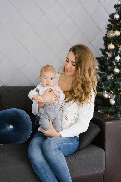 Portrait of a young mother in a white sweater and her young son. They're sitting on the couch by the Christmas tree - Foto, Imagen