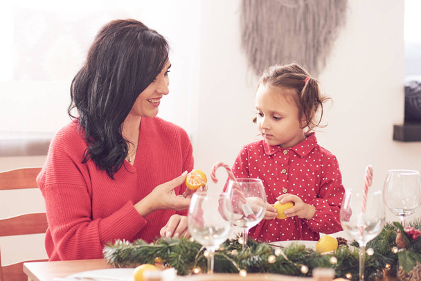 Iloinen äiti ja hänen tyttärensä nauravat, hauskaa joulukuusi ja tähden muotoinen evästeet keittiön pöydässä. Heillä on punaiset vaatteet, poronsarvet ja joulupukin hattupanta.
. - Valokuva, kuva