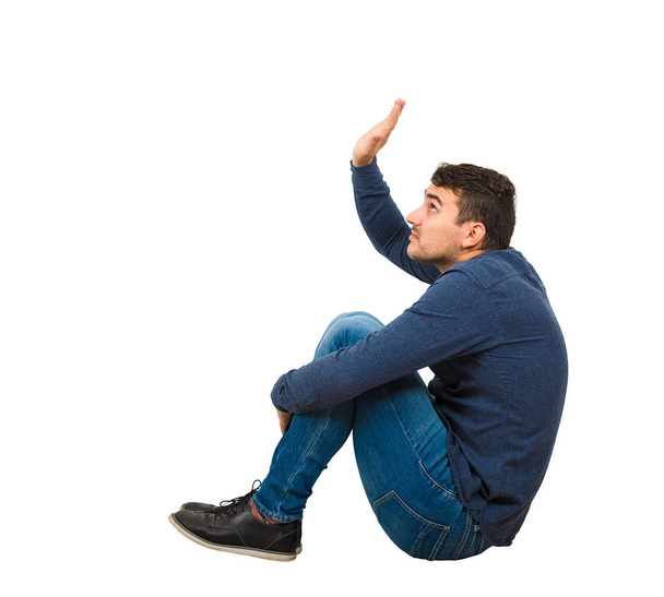 Πλευρική άποψη φοβάται νεαρός άνδρας εσωστρεφής κάθεται στο πάτωμα κρατά h - Φωτογραφία, εικόνα
