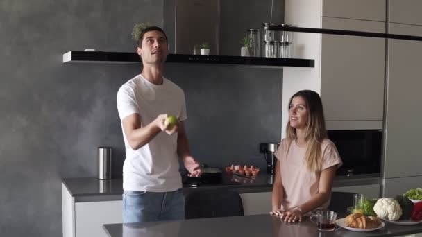 Attractive loving caucasian couple having fun in the home kitchen. Handsome man in jeans and white T shirt juggle with apples to impress his girlfriend, she hugs him. Slow motion - Video