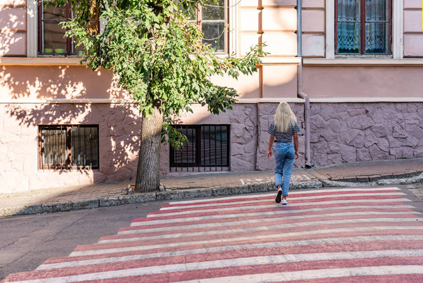 Summer sunny portrait of the attractive young blonde girl traveler in grey t-shirt and blue jeans on the red crosswalk among the old city center - Photo, Image