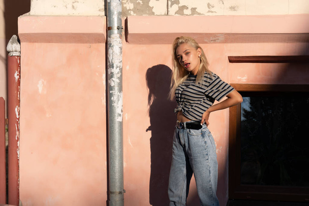 Fille blonde à la mode avec des cheveux lâches vêtus de t-shirt gris se tient au soleil près du mur rose de la vieille maison et tient la main sur sa taille
 - Photo, image