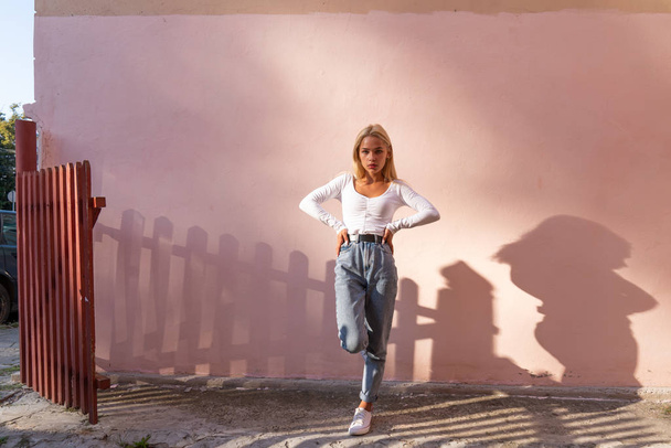 Модна блондинка в білому верхньому і синьому джинсах стоїть на одній нозі на сонці і дивиться на камеру, рожева стіна на фоні
 - Фото, зображення