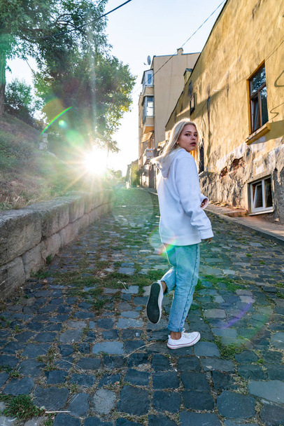 Retrato de moda de estilo callejero de la hermosa chica rubia con capucha blanca pasea por el camino de adoquines de la vieja ciudad europea, puesta de sol en el fondo
 - Foto, Imagen