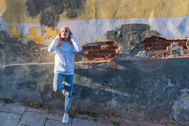 Attractive blonde girl in white hoodie leaned on the old grey wall and straightens her hair - Foto, afbeelding