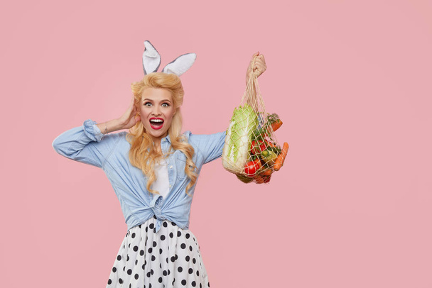 Happy Easter. Young pin up woman with bunny ears hold a string bag full of organic vegetables on pink background. Happy face. Emty space for text - 写真・画像