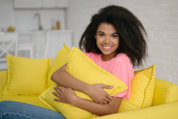 Young beautiful African American woman hugging pillow sitting on comfortable yellow sofa at living room. Authentic portrait of cute emotional curly haired girl relaxing and smiling at home - Photo, Image