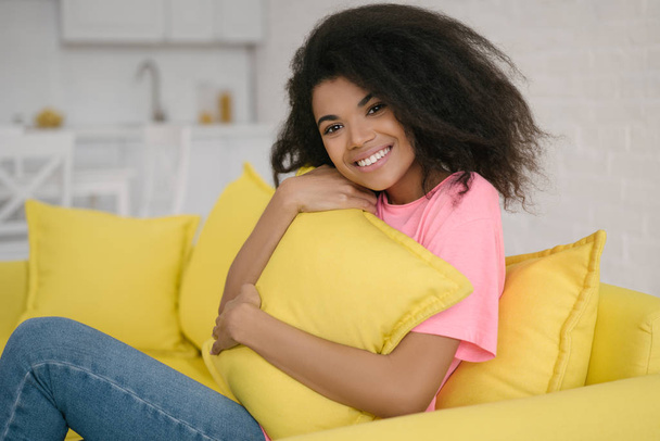 Authentic portrait of cute emotional curly haired girl relaxing and smiling at home. Young attractive African American woman hugging pillow sitting on comfortable yellow sofa at living room - Photo, Image