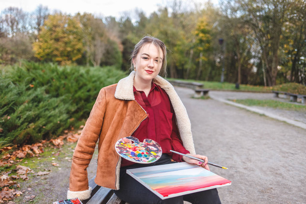 Retrato de uma artista de rua que se senta em um parque em um banco e segura uma pintura, paleta e escova em suas mãos
 - Foto, Imagem