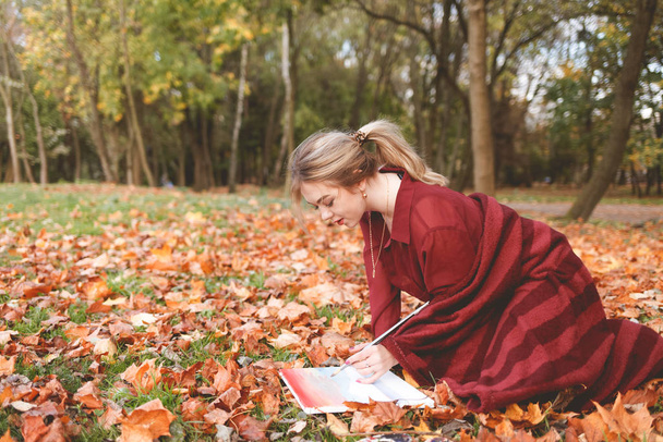 Nuori tyttö taiteilija piirtää kuvan polvillaan puistossa nurmikolla. Tyttö inspiroi luovuutta istuu syksyllä puistossa ja maalaus
 - Valokuva, kuva