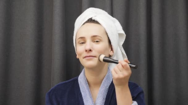 Smiling charming woman applying brush on face and looking at mirror.  Female doing makeup at home. Girl in blue bathrobe preparing for evening party and puts cosmetics on face - Felvétel, videó