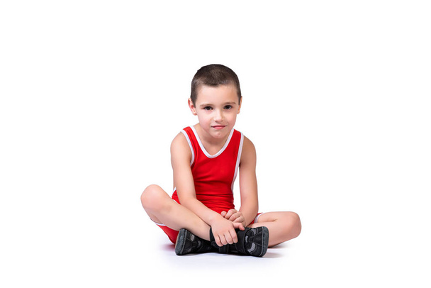 Menino alegre desportivo em uma meia-calça de luta azul está pronto para se envolver em exercícios esportivos, está sentado no chão em um fundo isolado branco
 - Foto, Imagem
