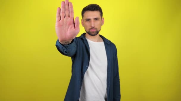 Young man in blue shirt and white T-shirt looks at camera, holds out his hand in front of him and says stop. Stand still. Yellow background with copy space. Stop action. 4k slowmotion footage. - Footage, Video