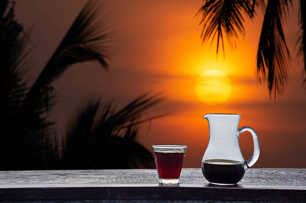 Una jarra y un vaso de café negro sobre una mesa de madera Fondo La luz dorada del sol y las nubes en el cielo con la sombra de los cocoteros
. - Foto, imagen