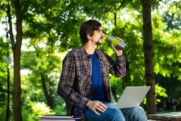 Молодой брюнет фрилансер с бородой и усами сидит с ноутбуком и кофе в парке
 - Фото, изображение