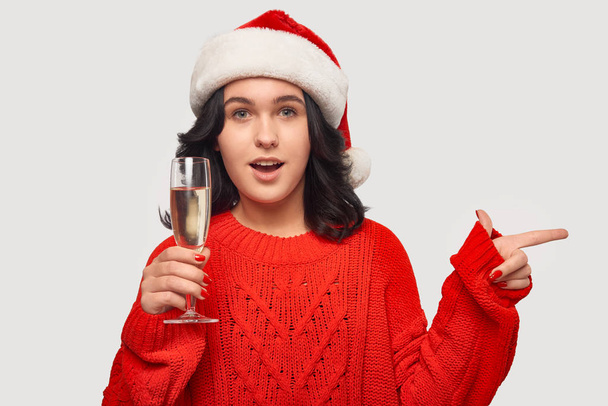 Girl in a red sweater and Santa hat holding glass of champagne and pointing at the copy space - Fotografie, Obrázek