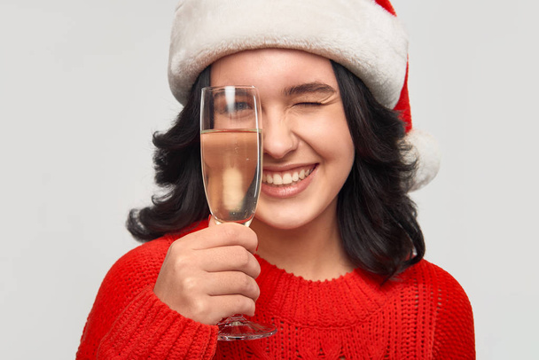 Hermosa chica morena en un suéter rojo y Santa mirando a través de una copa de champán
. - Foto, imagen