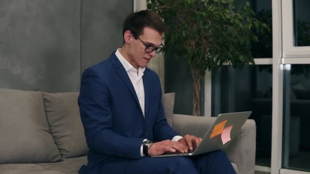 Caucasian businessman wearing formal blue suit and stylish glasses sitting on the sofa typing on his laptop on knees at the hotel room. Loft interior room - Footage, Video