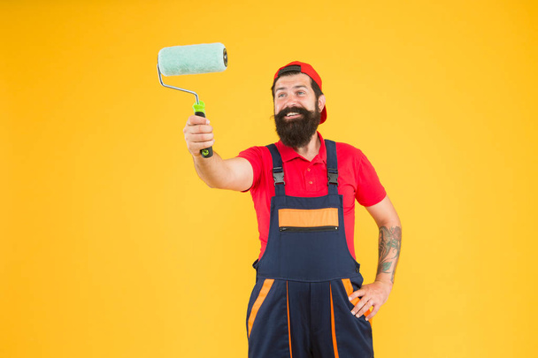 Room painting job. repair assistant in work uniform. engineer designer ready to paint wall. repair concept. happy bearded man worker with roller tool. hipster builder in cap yellow background - 写真・画像