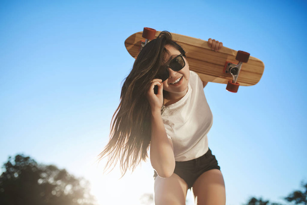 Sport, urban lifestyle and youth concept. Cheerful skater girl bending down to look camera, hold wooden penny board on shoulder, touch sunglasses, enjoy sunny summer holidays riding skateboard - Фото, изображение