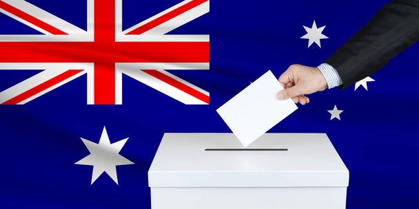 Εκλογές στην Αυστραλία. Το χέρι του ανθρώπου που βάζει την ψήφο του στην κάλπη. Κυματιστή σημαία Αυστραλίας στο παρασκήνιο. - Φωτογραφία, εικόνα