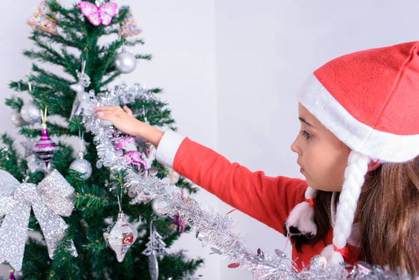 kleines Mädchen schmückt Weihnachtsbaum. Verkleidet als Weihnachtsmann. - Foto, Bild