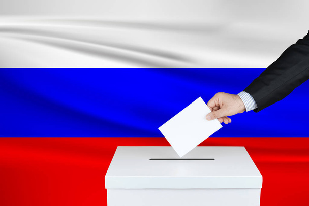Wahl in Russland. die Hand eines Mannes, der seine Stimme an die Wahlurne wirft. schwenkte die russische Flagge im Hintergrund.                                - Foto, Bild