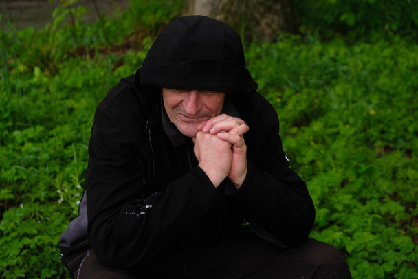 Чоловік у чорному капюшоні, який закрив очі. Чоловік з закритими пальцями дивиться вбік і посміхається. 58-річний чоловік у лісі чекає кінця дощу. Концепція самоізоляції
. - Фото, зображення