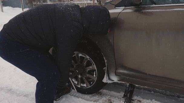 man raises car jack on road. man is changing wheel of car. replacement wheels in winter on road in a blizzard and snowfall. Breakdown of car, wheel change. - Photo, Image