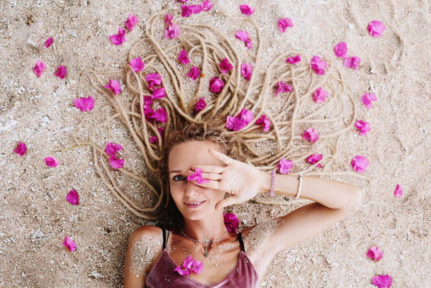 Young woman wearing dreadlocks hair style lying on sea shore with pink flowers in her hair - Foto, immagini