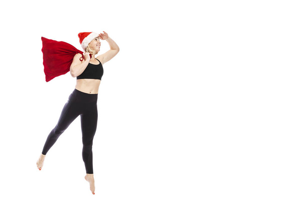 Young smiling woman in a black tracksuit in a Santa Claus hat and a red bag with gifts. Full height. Isolated over white background. Space for text. - Φωτογραφία, εικόνα