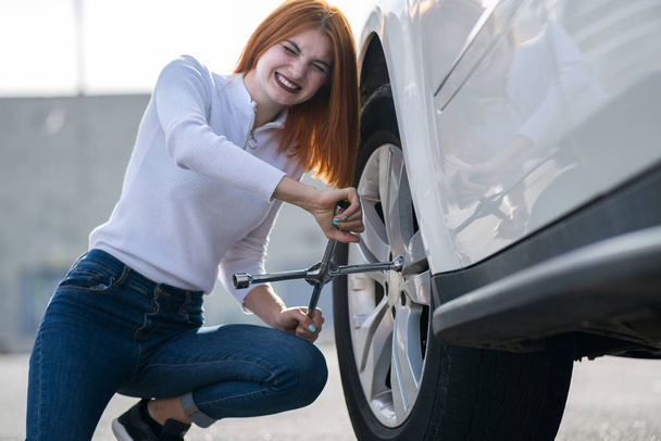 Młoda kobieta z kluczem zmieniającym koło na zepsutym samochodzie. - Zdjęcie, obraz