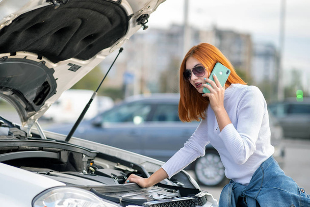 Young woman standing near broken car with popped hood talking on - Photo, image