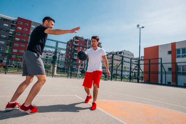 Zwei kaukasische Freunde tragen rote Sportgeräte und spielen an einem sonnigen Tag im Freien Basketbälle, während sie dribbeln und sich gegenseitig pushen. - Foto, Bild