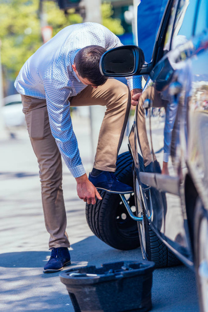 Un joven empresario cansado sudoroso se limpia el sudor de la frente mientras intenta cambiar un neumático pinchado en un hermoso día soleado. - Foto, Imagen